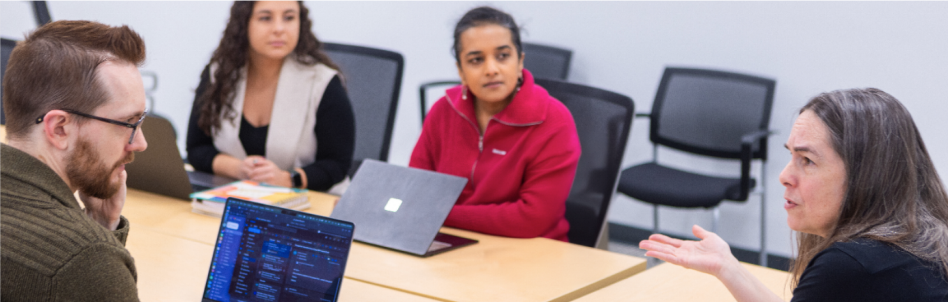 Four people sit around a conference table engaged in discussion. One woman gestures as she speaks while others listen, with laptops and notebooks open in front of them. The setting appears to be a classroom or meeting room.