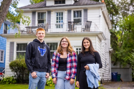 Three students stand in front of a house. 