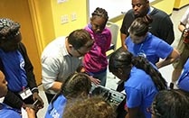 Group of students standing around a teacher showing them a computer component. 