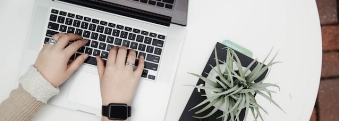 A circular white table with a laptop computer. am aloe plant, a cell phone, two pencils, a pair of cat eye glasses, and a planner. There are hands typing on the laptop computer.