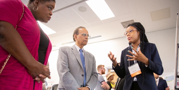 An undergraduate student in business apparel and a name tag discusses their research with two individuals, using their hands to communicate expressively.