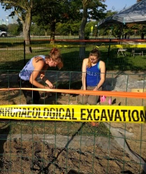 Two people doing archaeological work with a yellow tape that says ARCHAEOLOGICAL EXCAVATION in front of them.