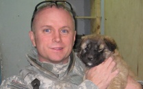 Portrait of William Mandrick, PhD, on a deployment, with a puppy found after a firefight with al Qaeda insurgents. Puppy was adopted by US forces. Mandrick observes, "We liked having dogs around because they sense when something is not right.". 