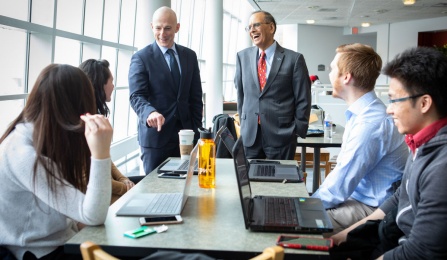 A group of students at a table look toward two people standing.