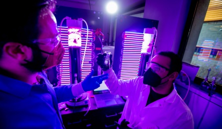 Two researchers look at an oject in a room with colorful lighted tubes.