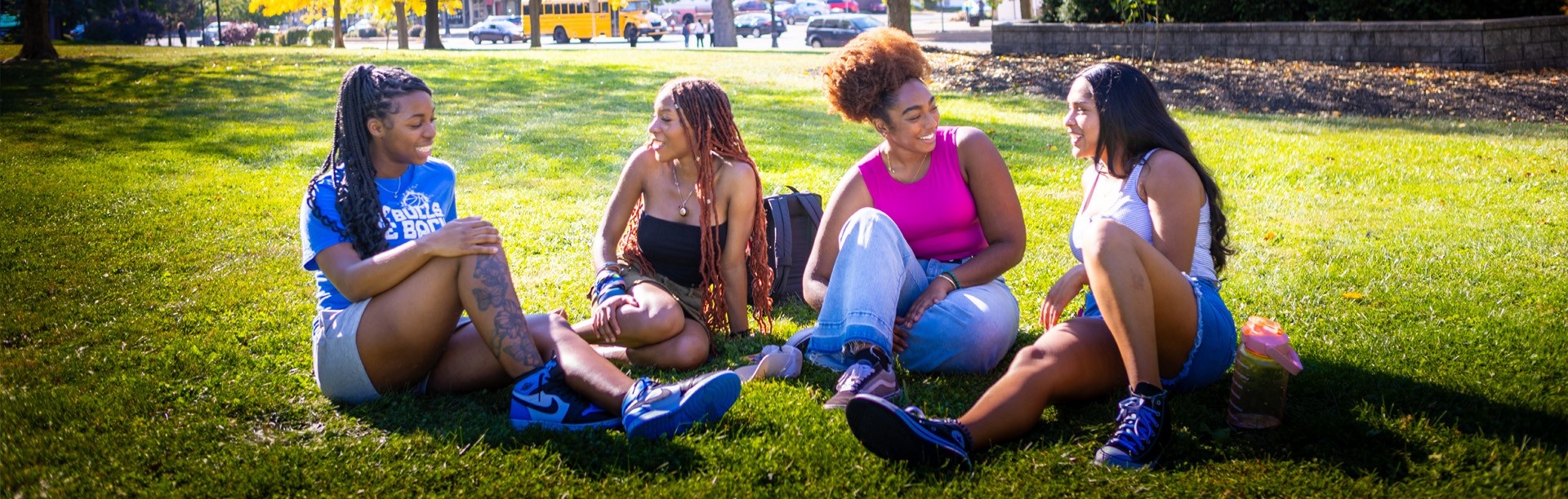 UB students sitting on the grass on campus in the sun, laughing and talking together. 