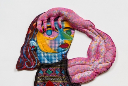 a textile wall work by Maria Guzman Capron. The bust of a figure, made of lightly stuffed fabric and paint, looks out towards the viewer, her hand on her head. 