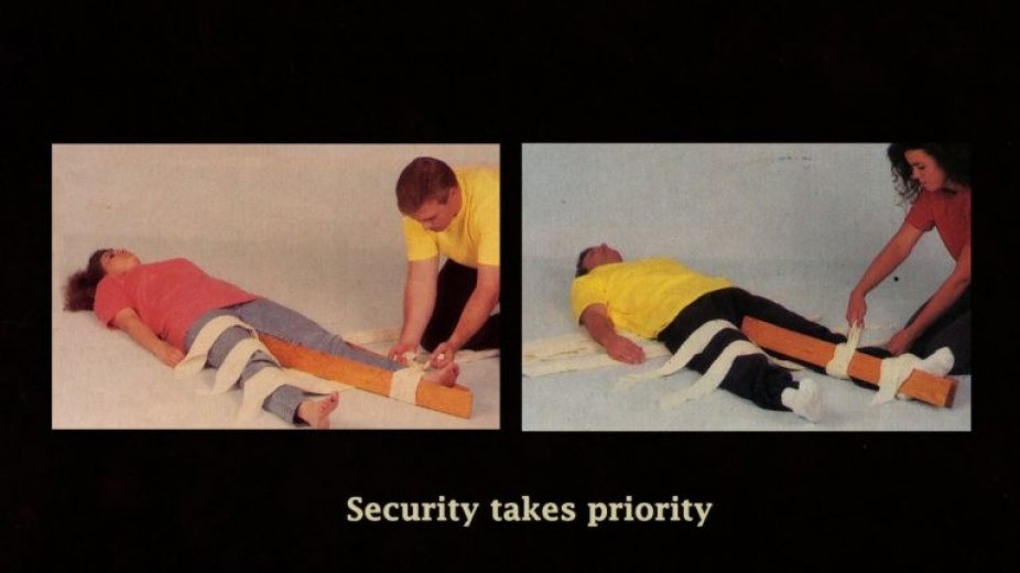 Sill Image from Video by Kelly Sears juxtaposing two images; woman with pink shirt lying on the floor and the man on the right applying a bandage on woman's left leg with wooden stick(left image), man with yellow shirt lying on the floor with a woman on the right applying a bandage with wooden stick on the man's left leg(right image) and 'Security takes priority' on the bottom center of the still.
