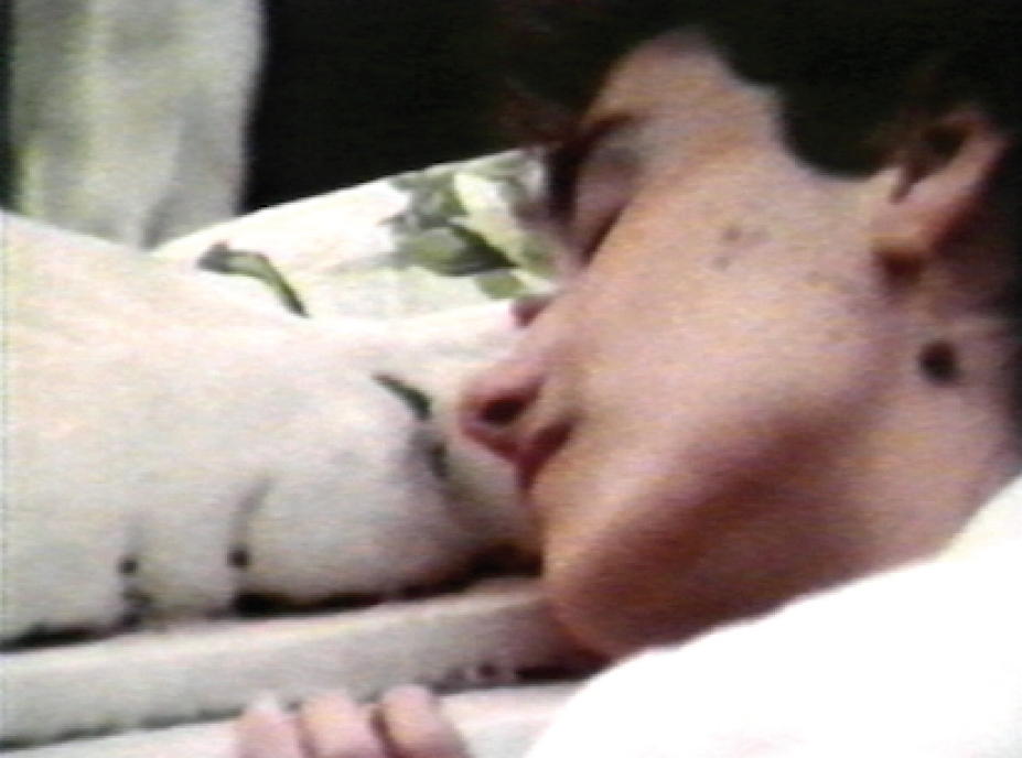 Still Image from Caterina Borelli's video of a figure lying in profile on white and green comforter.