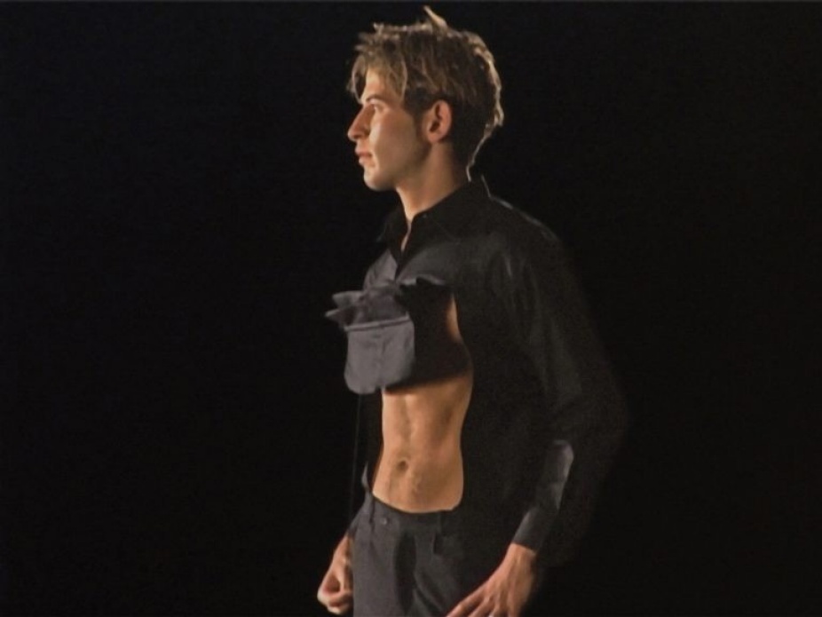 A photo of a man in profile. He wears a black shirt that is pulled up in front, revealing very tight abs.