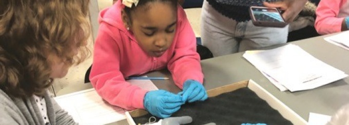 A student from Bennett Park Montessori holding and UB Museum Studies students handling artifacts as a teacher looks on.