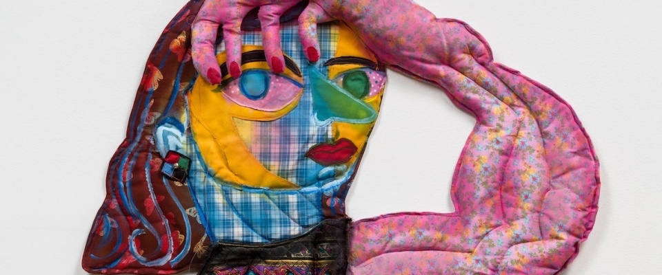 a textile wall work by Maria Guzman Capron. The bust of a figure, made of lightly stuffed fabric and paint, looks out towards the viewer, her hand on her head. 