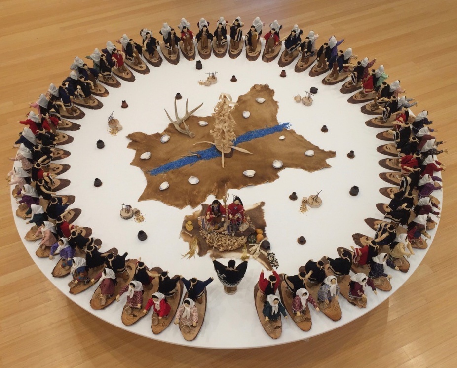 A photo of a work by Elizabeth Doxtater. On a white round table a circle of corn husk dolls in boats faces outward. In the center of the circle is a cow hide and more corhnusk dolls arranged in ceremony.