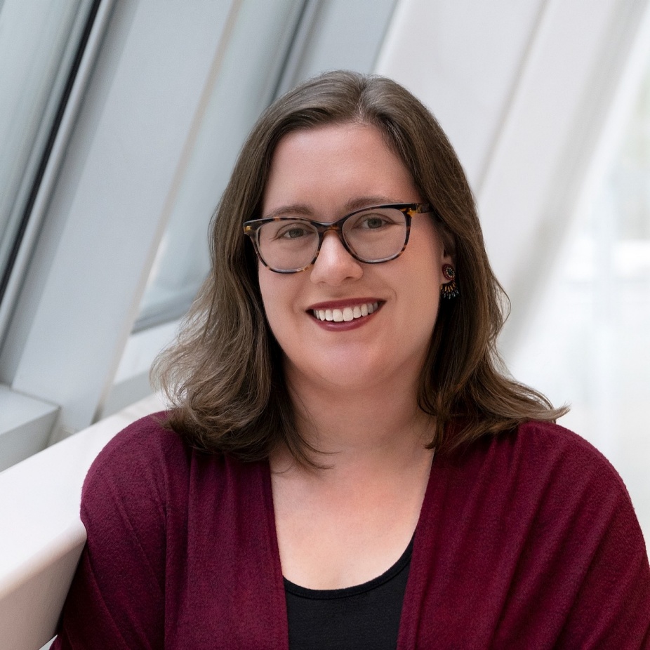 A photograph of Anna Wager, a smiling woman with light brown hair wearing glasses and a red sweater.