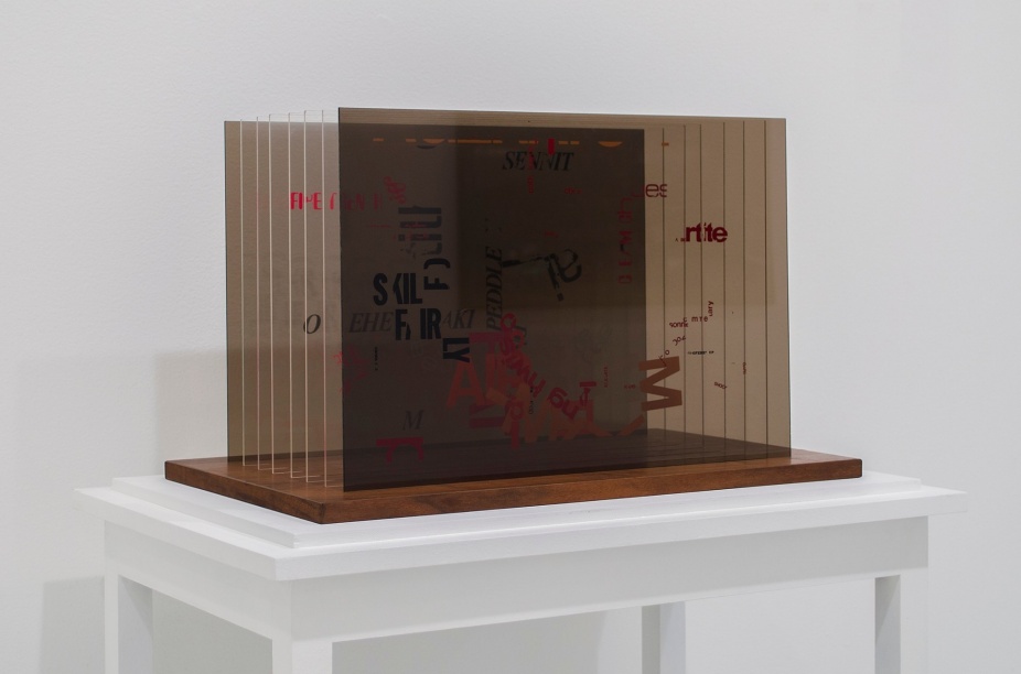 A photo of a John Cage sculpture where 8 plexiglass panels have silkscreened letters in a random and anarchic order.