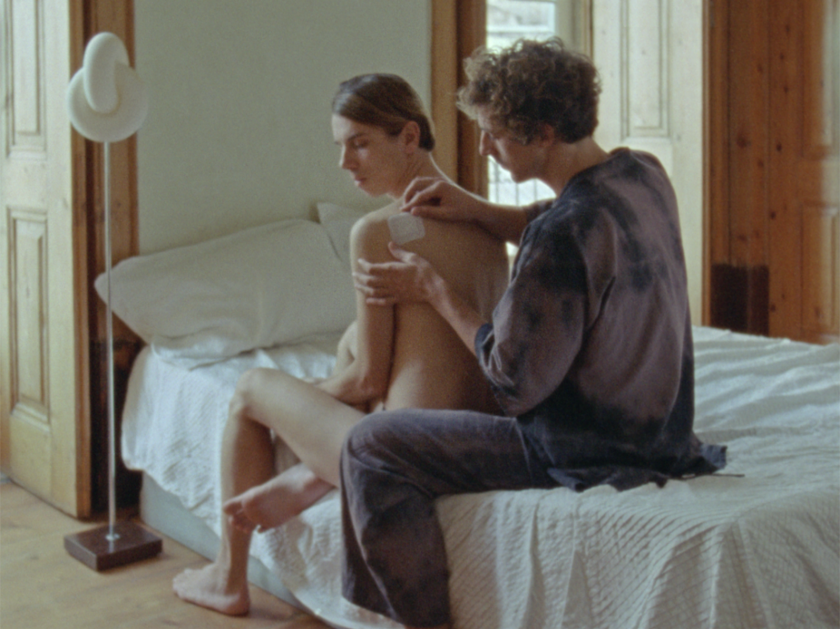 A person wearing a white tank top stands amongst household, cosmetic and pharmaceutical items dangling from strings. This is a still from Every Ocean Hughes’s video entitled One Big Bag.