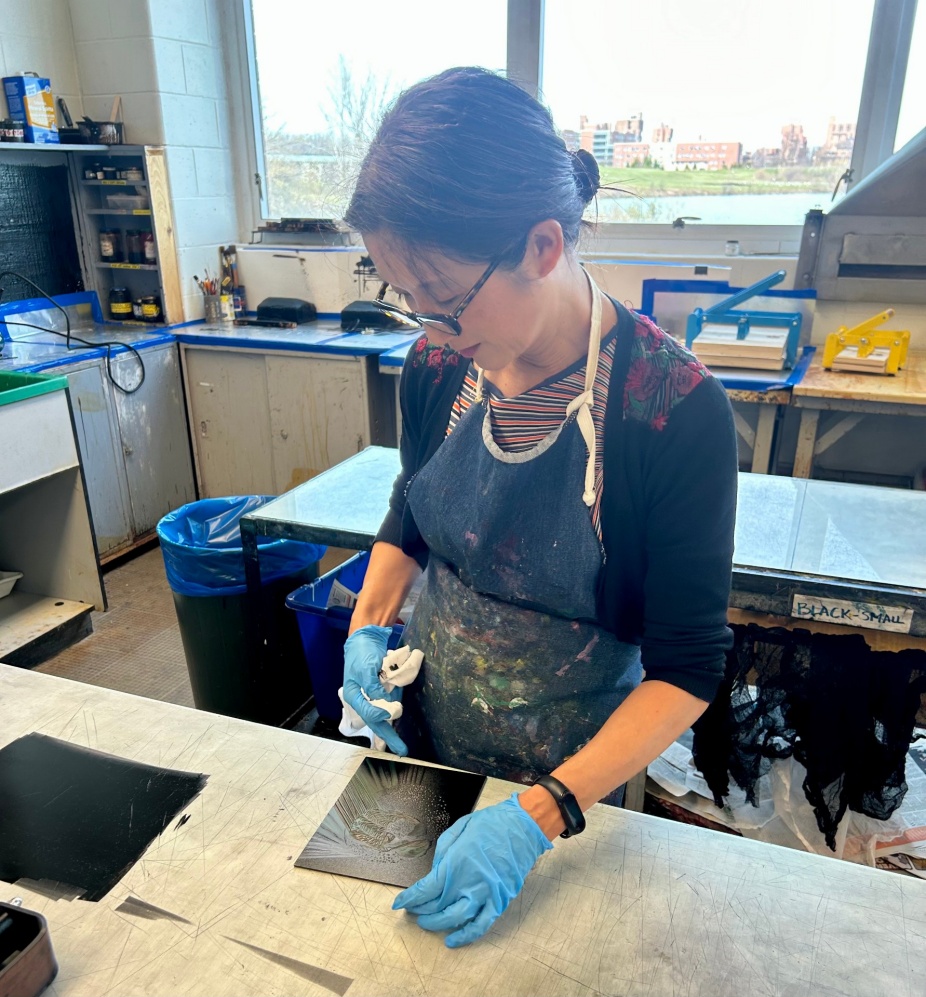 a photo of artist Wuon-Gean Ho, an Asian woman, sitting in a park and drawing in a sketch book. She has a yellow backpack and yellow waterbottle next to her, and is wearing a hat, and brightly patterened sweater.
