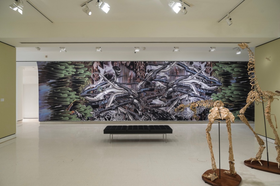 A documentation photograph of the exhibition Christy Rupp: Runoff where two tall bird-like skeletons are in the foreground and a large mural of pipelines is in the background. 