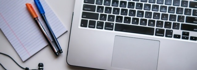 A laptop computer. To the left of the computer is a pair of earbud headphones, a notepad, and two pens - an orange pen and a blue pen. Photo Credit: Maya Maceka. 