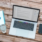 A laptop open on a wooden table. To the left of the computer is a notepad with a pen on top and a glass of water. To the right of the computer is a smartphone and a potted plant. 