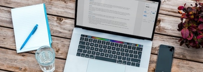 A laptop open on a wooden table. To the left of the computer is a notepad with a pen on top and a glass of water. To the right of the computer is a smartphone and a potted plant. 