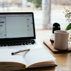 A laptop open on a wooden table. In front of the computer is a notepad with a pen on top. To the right of the computer is a pot of tea, a teacup, and a potted plant. 