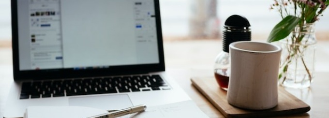 A laptop open on a wooden table. To the left of the computer is a notepad with a pen on top and a glass of water. To the right of the computer is a smartphone and a potted plant. 