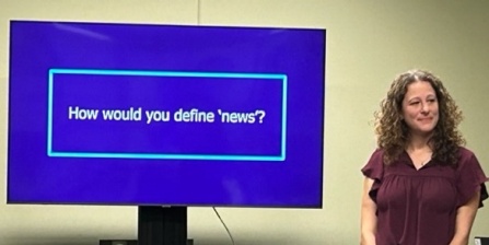 Image of presenter in front of a screen reading "How would you define 'news'".