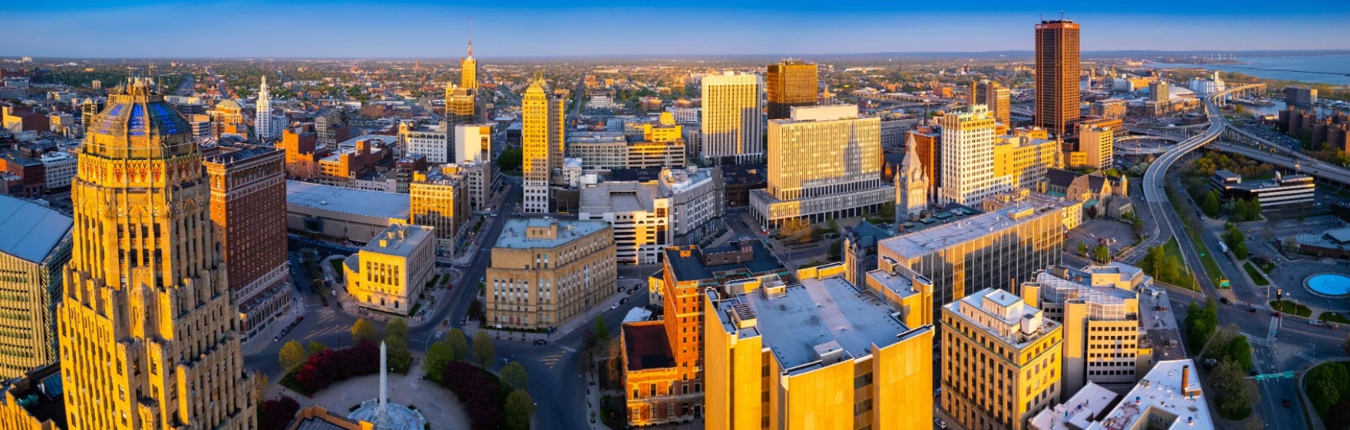 Birdseye aerial view of the City of Buffalo at sunset. The expansive urban scene faces south east. In the foreground, highlighted by the golden light of sunset, are several buildings in the downtown district, including the art deco style City Hall, and the ultra modern Court House. In the background, residential districts are visible in the distance. In the upper right corner, the Skyway winds out toward the shoreline of Lake Erie, showing the windmills of Lakawanna. The Boston hills appear in the distance.