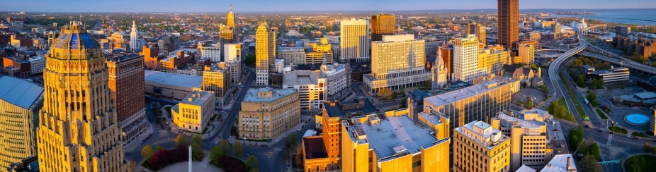 Birdseye aerial view of the City of Buffalo at sunset. The expansive urban scene faces south east. In the foreground, highlighted by the golden light of sunset, are several buildings in the downtown district, including the art deco style City Hall, and the ultra modern Court House. In the background, residential districts are visible in the distance. In the upper right corner, the Skyway winds out toward the shoreline of Lake Erie, showing the windmills of Lakawanna. The Boston hills appear in the distance. 