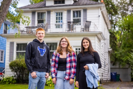 Three students stand in front of a house.