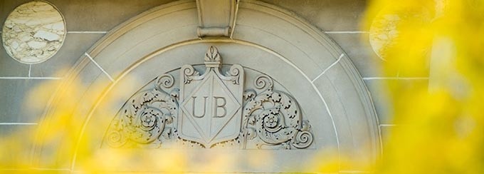 Photo of UB emblem on Foster Hall, with blurred yellow leaves framing the photo, in the foreground.