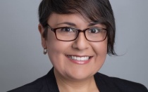 Photo shows a woman with dark hair wearing glasses and a black blazer, smiling at the camera. 