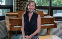 Person with a black vest, long hair, earring, sitting on a piano bench. 