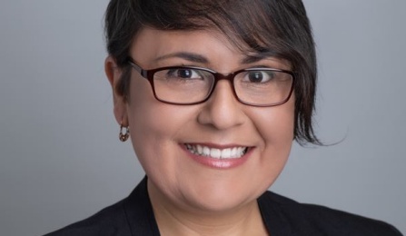 Photo shows a woman with short dark hair, wearing glasses and a black blazer, smiling at the camera. 