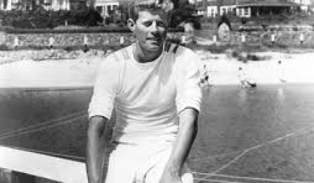 JFK, Jr wearing all white t-shirt and short sitting on a railing with a lake and houses behind him. 