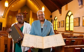 Two women within a church holding documents, smiling at the camera.