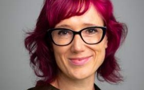 Photo shows a woman with pink hair and black glasses, smiling at the camera.