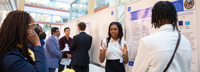 Photo shows a student discussing their research poster to a group of people. 