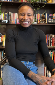 Image of a woman with dark hair, smiling at the camera. They are sitting in front of a bookshelf, wearing a black turtleneck shirt and jeans. 