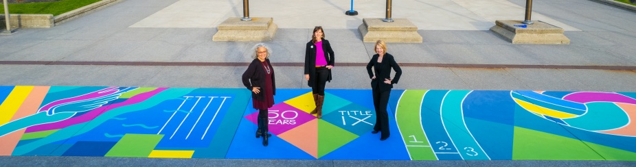 UB female student-athletes pose together by the new Title IX mural outside Alumni Arena in October 2022 for an aerial image. From left, Gender Institute Director Carrie Tirado Bramen, mural artist Cassandra Ott and Contemplative Sites Subcommittee chair Kelly Hayes McAlonie.