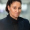 A woman wearing black high neck neck shirt with blue blazer, looking at the camera. 