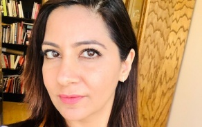 Photo shows a woman with dark hair and a blue and black striped shirt in front of a book shelf.