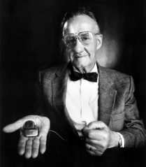 in black-and-white of a silver-haired man in a coat and bowtie holding out a medical device in his palm.