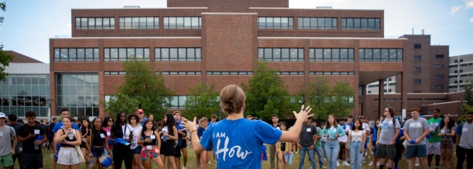 person in "I am how" shirt addressing a group of students with Capen Hall in background. 