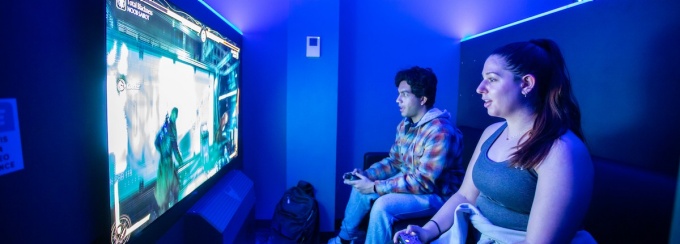 two students in a dark room playing a video game on a big screen. 