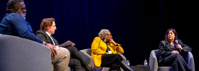 Four panelists sit on stage in gray armchairs during a discussion event. From left to right, a man in a blue shirt, a man in a suit holding papers, a woman in a bright yellow blazer and black boots, and a woman in a dark suit holding a microphone while speaking. They are positioned under a large blue screen on a dark stage, with water bottles on a small table between them.