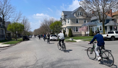 Approximately 10 individuals ride bikes through an urban neighborhood. It is a sunny day.