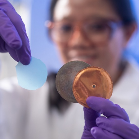 A laboratory researcher wearing protective goggles and a white lab coat holds up a light blue circular hydrogel sample with purple nitrile gloves. In their other hand, they display what appears to be a copper or copper-colored testing apparatus or holder with a mesh or filter component. The image is focused on the materials being held, while the researcher appears blurred in the background, smiling.