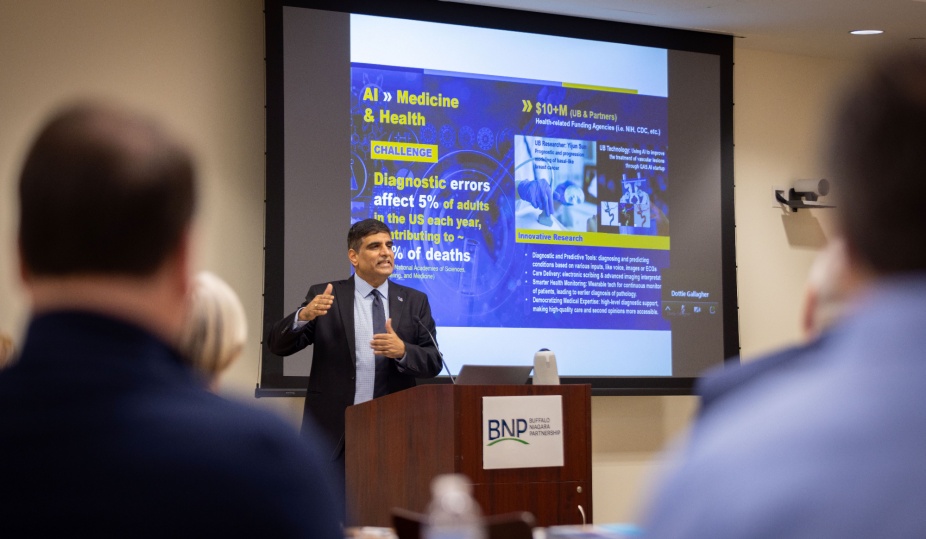 Dr. Venu Govindaraju stands at a podium, speaking to an audience. Behind him, a slide entitled AI, Medicine & Health can be seen.